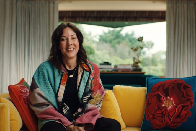 Leslie Blodgett sits on a vibrant yellow couch, wearing a colourful jacket, with a bright window, greenery, and decorative pillows in the background.