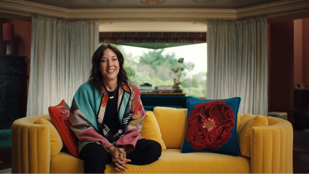 Leslie Blodgett sits on a vibrant yellow couch, wearing a colorful jacket, with a bright window, greenery, and decorative pillows in the background.