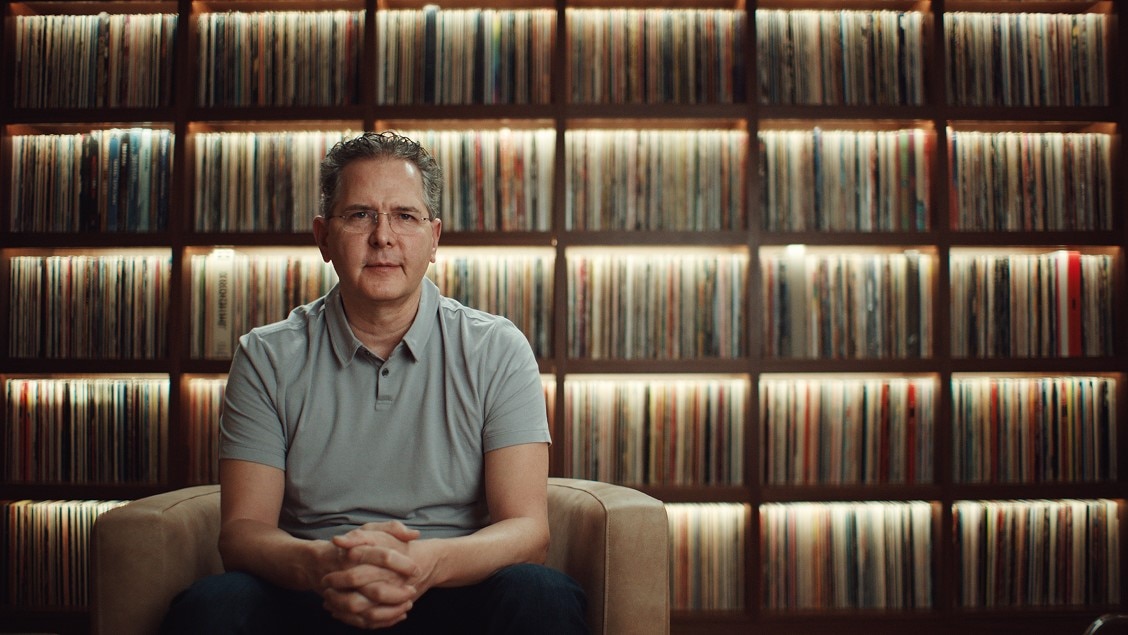 Craig Kallman sits in a beige armchair, wearing a light gray polo shirt and dark jeans. He is centered in front of a large, warmly lit wall of vinyl records arranged in wooden shelves, creating a cozy and nostalgic atmosphere.