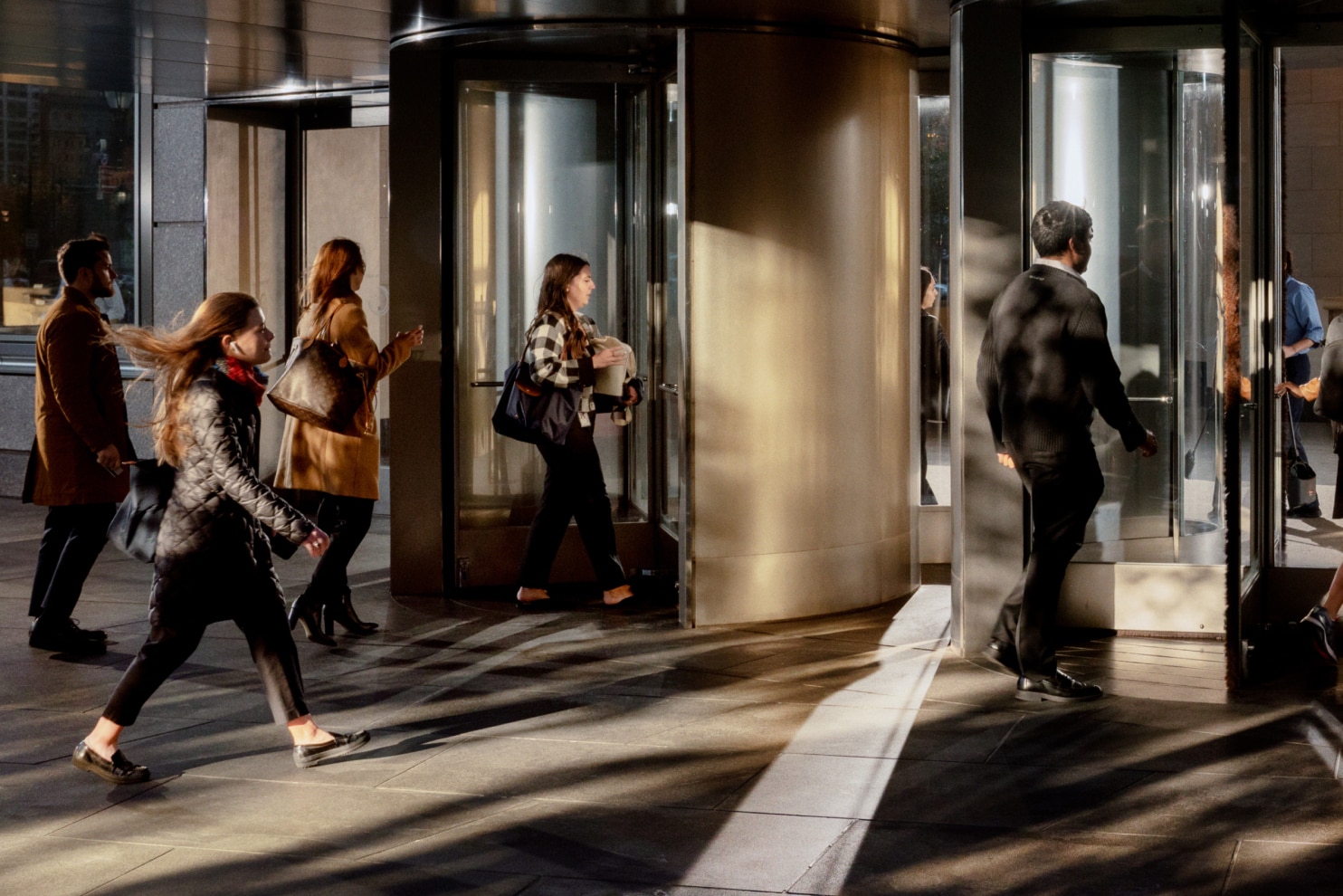 Three professionally dressed individuals walk through a stylish, modern lobby with curved architectural elements. The man on the left, wearing a green jumper and dark trousers, carries a brown leather duffle bag. The woman in the middle wears a striped blouse with a jumper draped over her shoulders and carries a black handbag. The woman on the right, dressed in a navy blue top and wide-leg trousers, pulls a sleek silver rolling suitcase while holding a phone. They appear engaged in a friendly conversation in a travel or business setting.