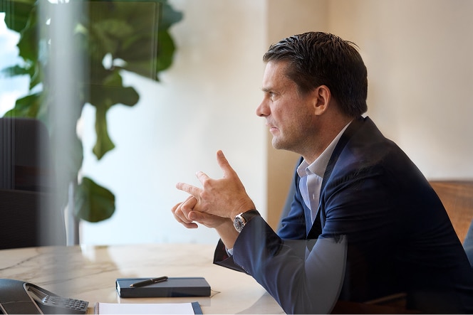 A man in a suit, sitting at a table, clasps his hands. A pen, planner, and potted plant are on the table, with framed pictures in the background.