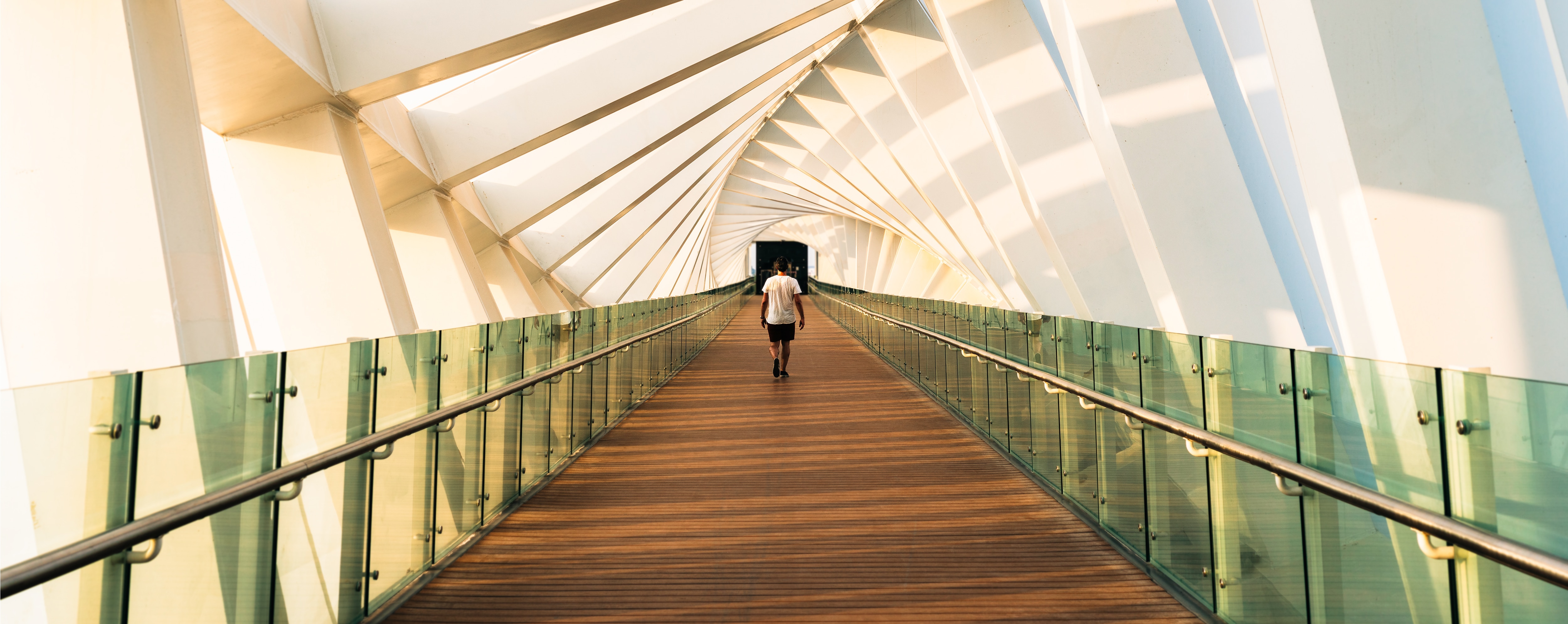A person walks along a modern, enclosed pedestrian bridge with geometric white beams and glass railings. Sunlight casts angular shadows on the walls, creating a dynamic pattern