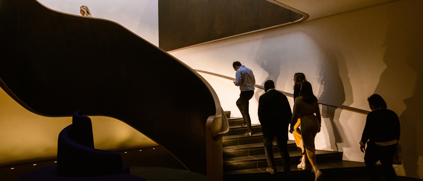 A group of people walks up a dimly lit, modern staircase with curved railings. Warm lighting casts dramatic shadows on the walls