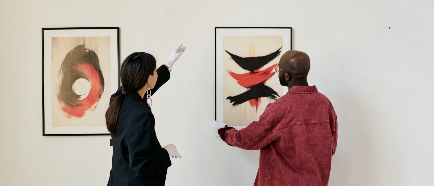Two people view and discuss abstract art in a gallery, with one pointing toward a framed painting featuring bold red and black brushstrokes.
