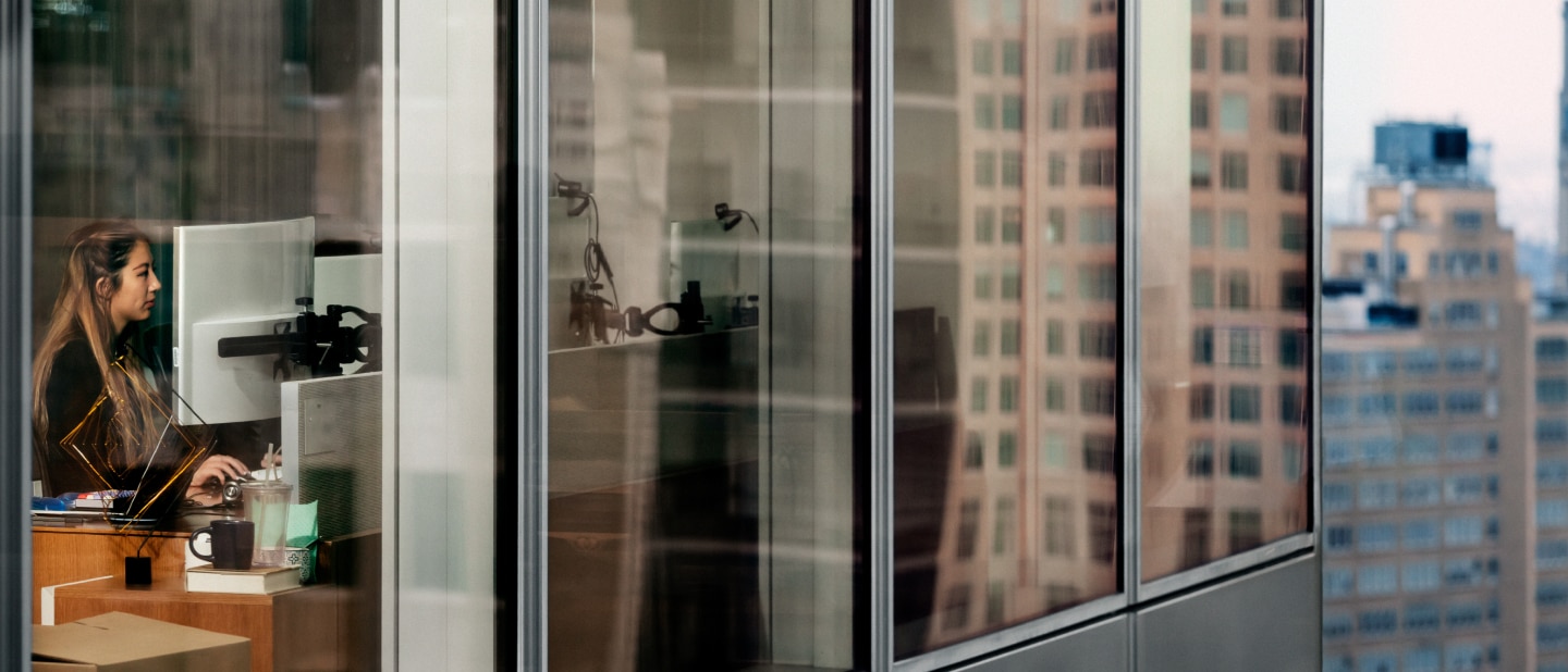 “A woman works at her desk in a modern high-rise office, seen through large glass windows reflecting the surrounding city buildings. The scene captures a blend of interior workspace and urban skyline ”