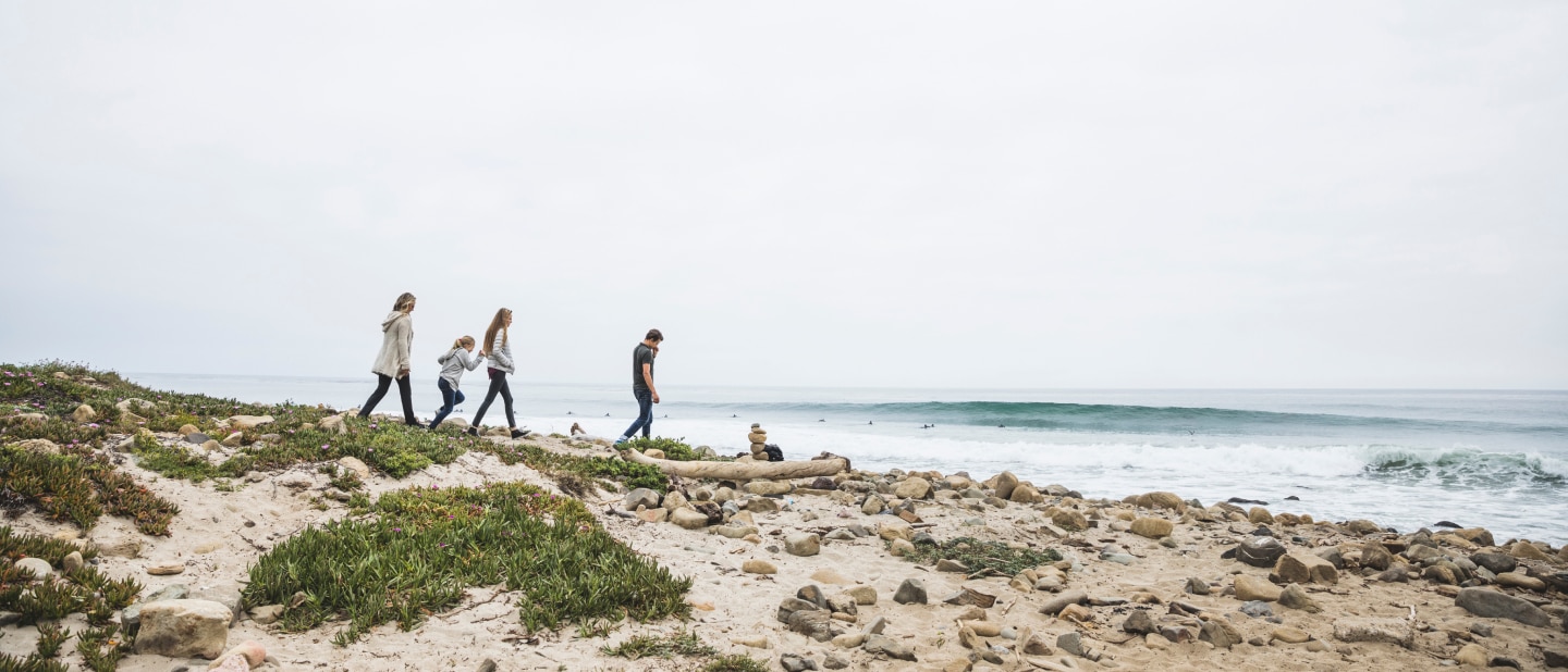 A family walks along a rocky beach path near the ocean, surrounded by sand, greenery, and scattered stones. Waves roll in as surfers are seen in the distance under an overcast sky
