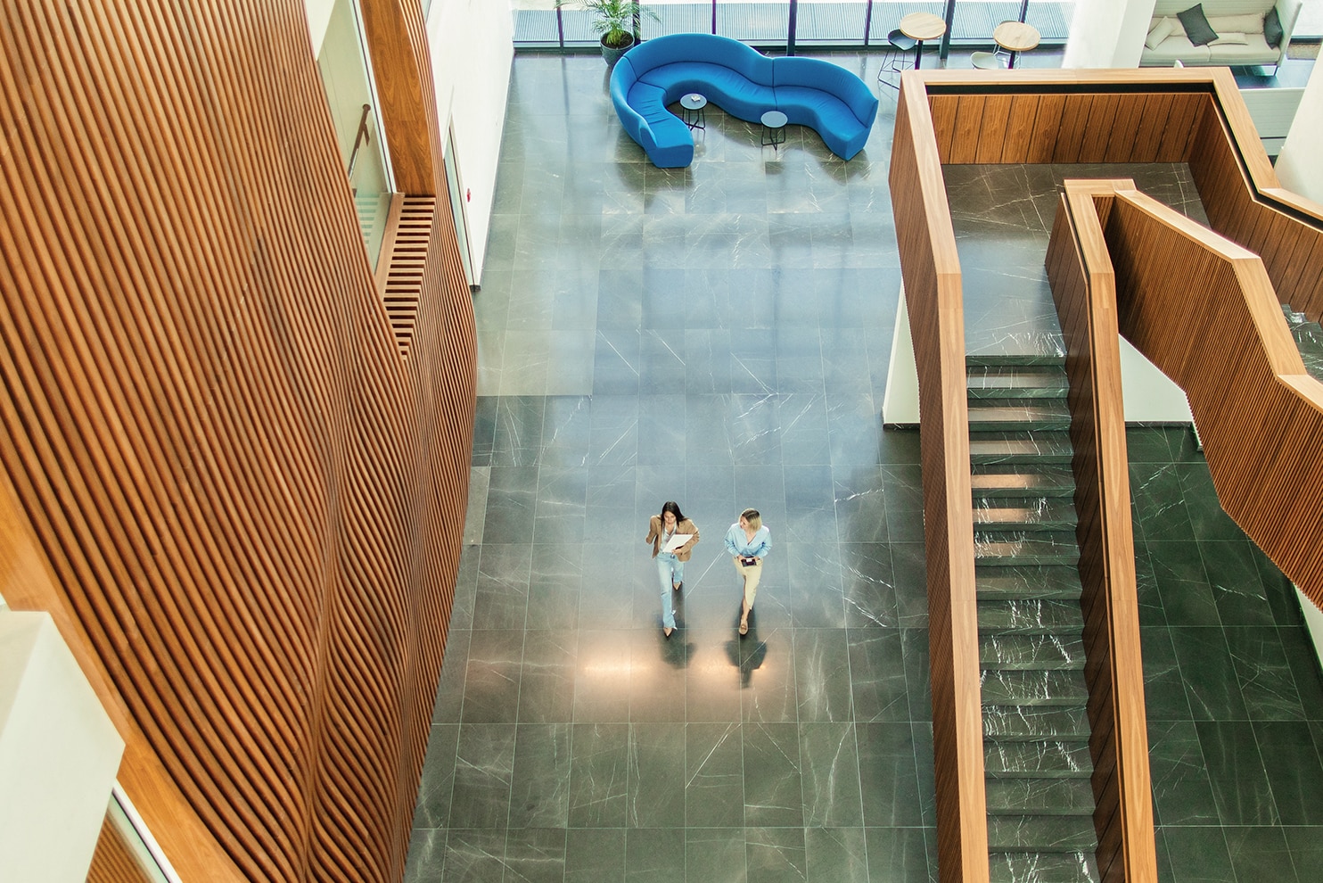 Two people walk through a spacious modern atrium with high ceilings, wooden panel walls, and a geometric staircase. A curved blue seating area is visible in the background