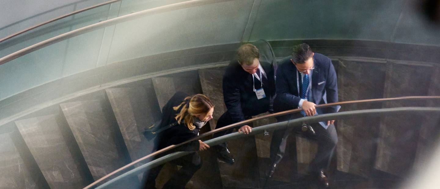 Three professionally dressed individuals walk down a curved glass staircase, engaged in conversation. They wear name badges, suggesting a conference or business event setting.
