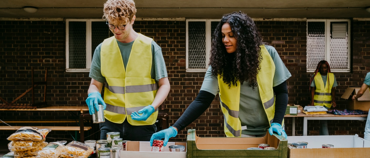 Two volunteers wearing reflective vests and gloves sorting canned goods at an outdoor food distribution table.