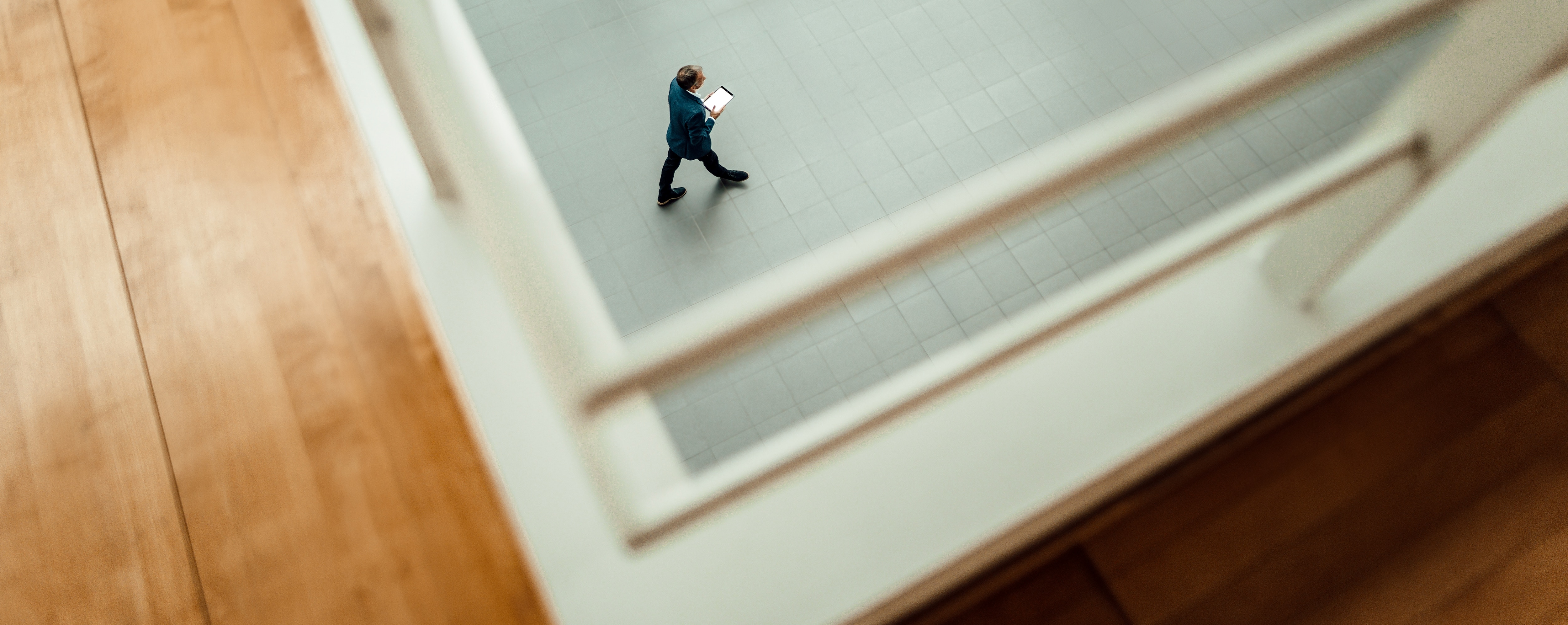 A man in a blue blazer walks across a tiled floor, holding a tablet, viewed from above through a railing. The perspective creates a framed effect, emphasising movement and space