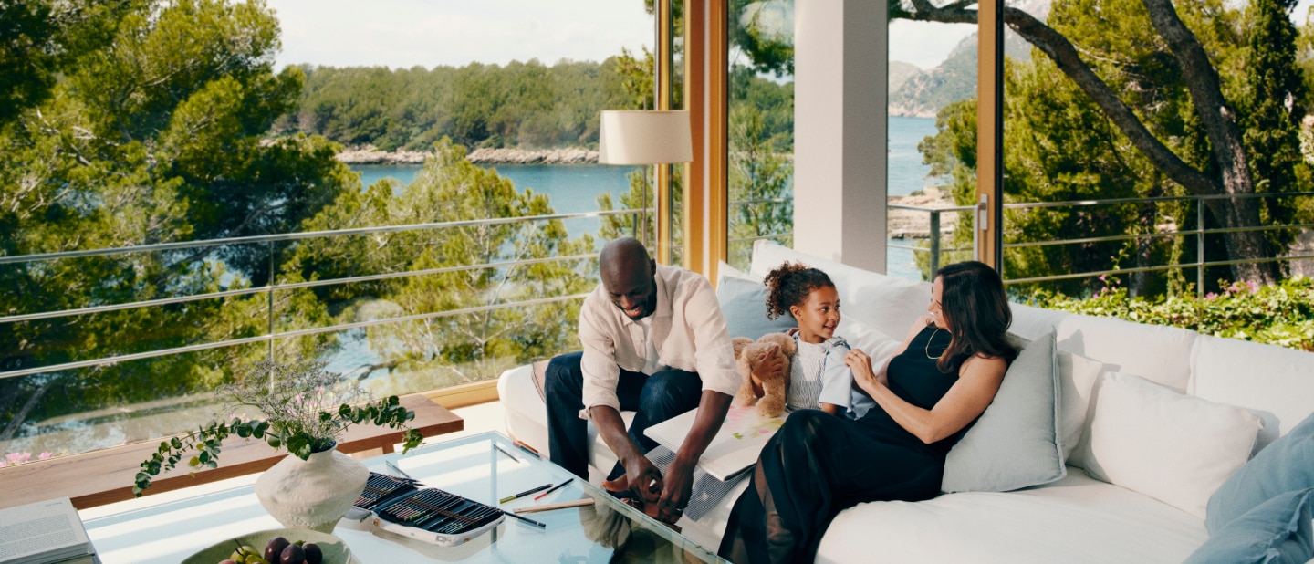 A family relaxes in a bright, modern living room with floor-to-ceiling windows overlooking a scenic coastal view. A man and a young girl are drawing together while a woman watches, smiling. The space is filled with natural light, greenery, and cosy furnishings.