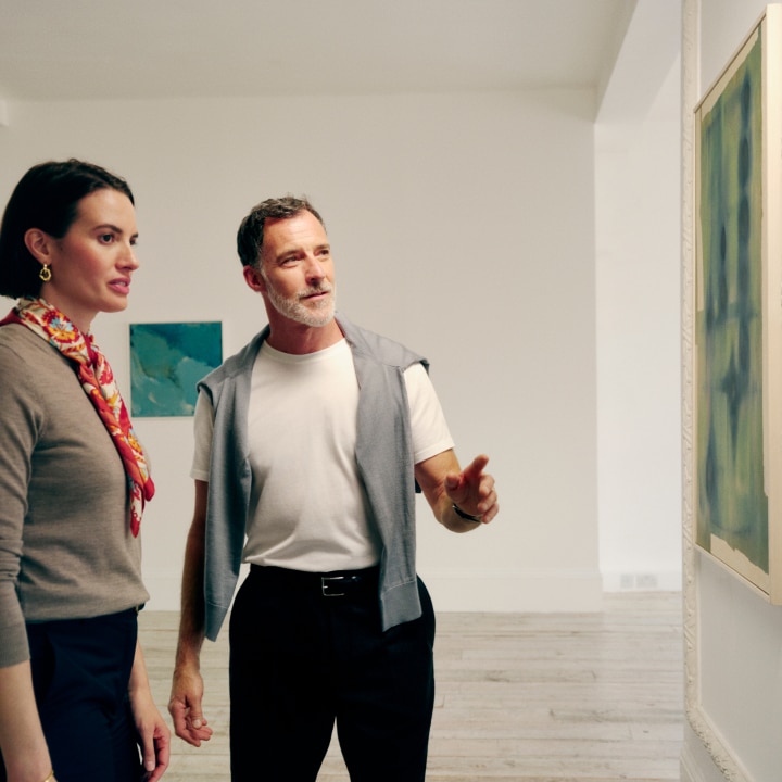 A man and a woman stand in an art gallery, engaged in conversation while observing a piece of artwork on the wall. The man gestures toward the artwork as he speaks, and the woman listens attentively. The gallery setting is minimalist, with white walls and framed abstract paintings