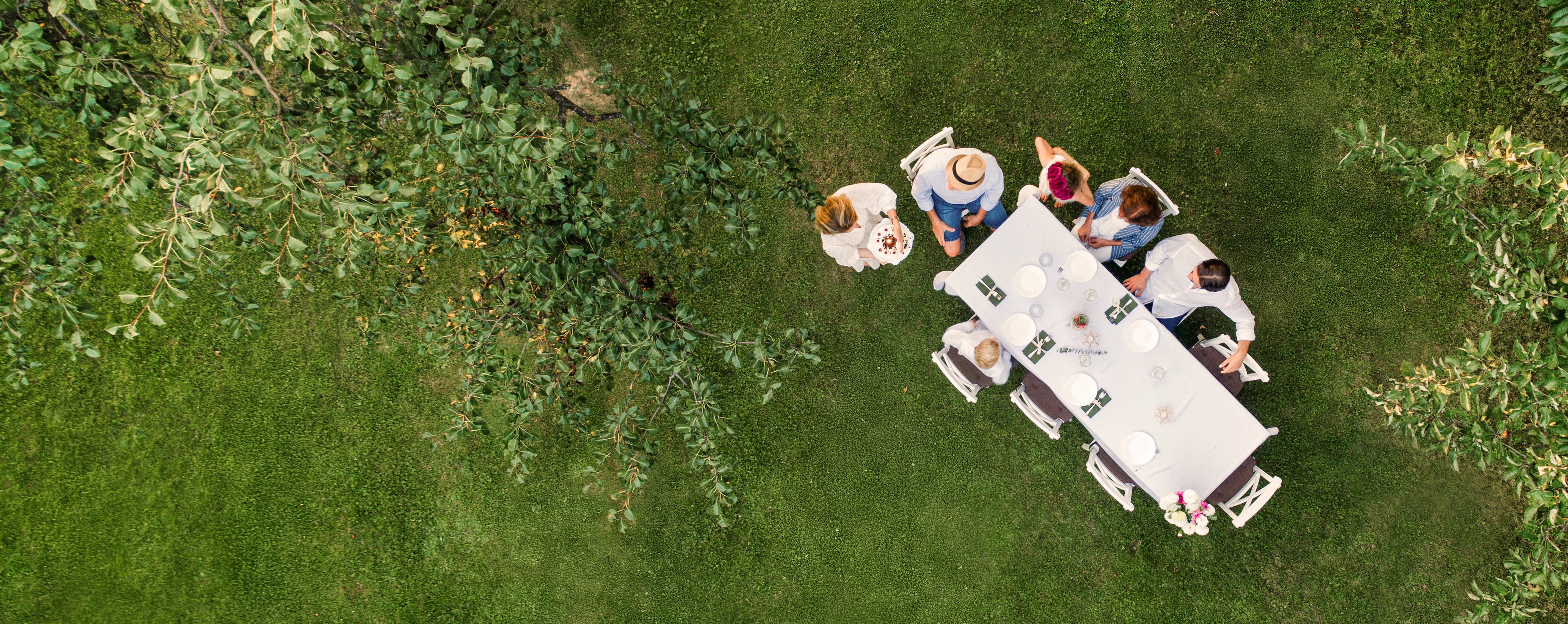 A group of people gathers around a long, white-clothed table set for a meal in a lush green garden. The aerial view captures the setting among trees